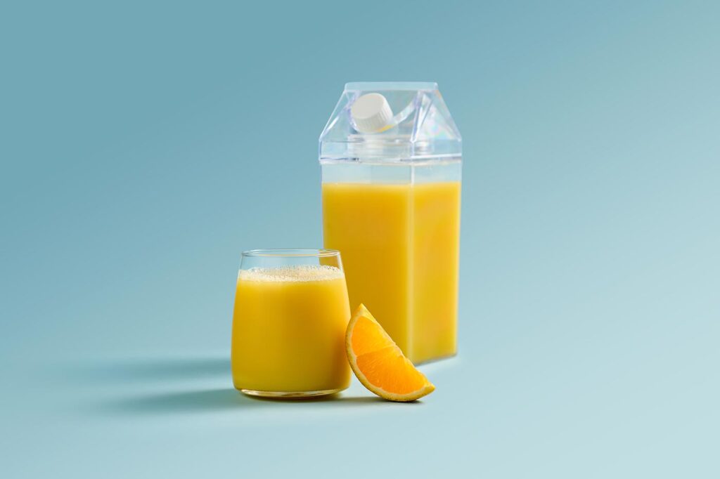 A clear container of Florida Orange Juice with a glass of OJ and an orange wedge next to it