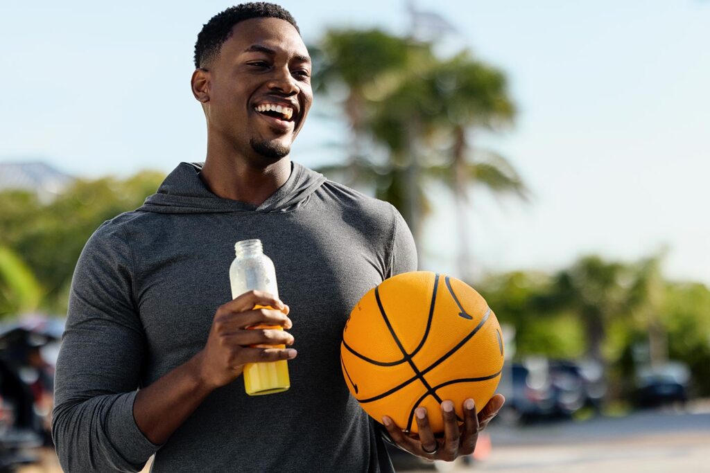 A smiling man standing outside at a park holding a basketball with a clear bottle of Florida Orange Juice